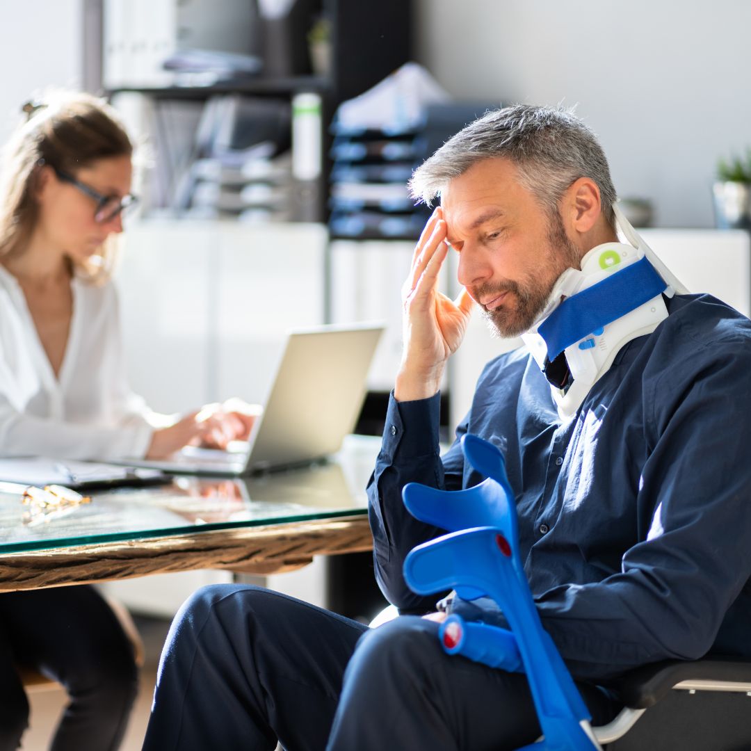 An injured man sits in an office holding crutches and wearing a neck brace, appearing concerned while a woman works on a laptop nearby. This image represents the aftermath of a pedestrian accident and the legal consultation process related to PIP (Personal Injury Protection) coverage in Washington, DC. Regan Zambri Long PLLC assists clients in navigating PIP claims for medical bills and lost wages following pedestrian incidents.