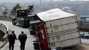 A large semi-truck lies overturned on a curved highway ramp in Fredericksburg, Virginia, as two uniformed officers inspect the crash scene while a tow truck prepares recovery. This incident represents the type of catastrophic collisions handled by truck accident lawyers at Regan Zambri Long PLLC. These accidents often involve major injuries, insurance disputes, and complex liability, requiring experienced legal help. Victims of truck crashes in Fredericksburg can contact the firm at +1 (202) 937-3310 to pursue compensation and protect their rights.