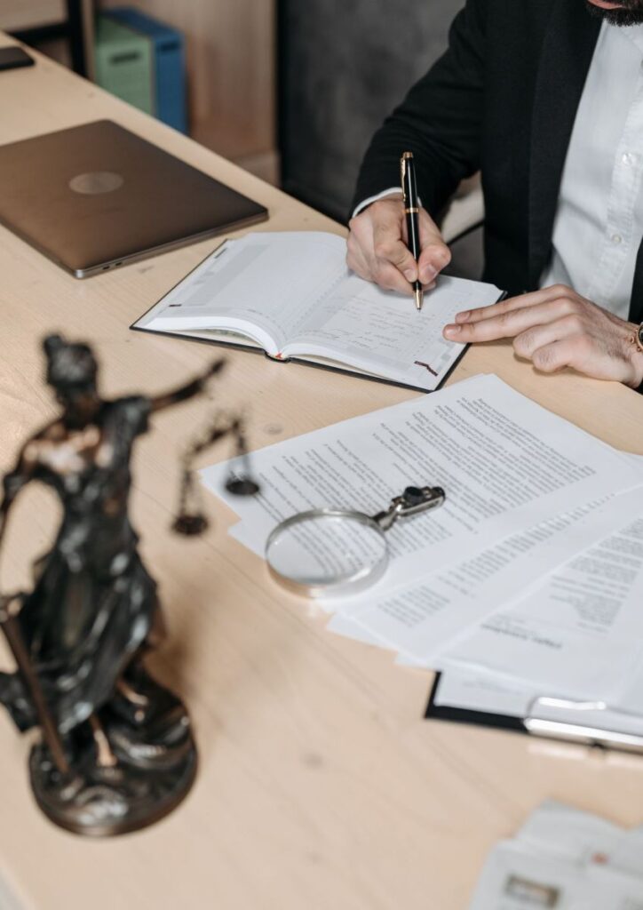 A Prince George’s County, MD personal injury lawyer works at a wooden office desk with legal documents, a pen, and a magnifying glass in view. The attorney is writing notes in a case planner while reviewing legal paperwork, and the Lady Justice statue appears in the foreground. This scene reflects the meticulous preparation and dedication lawyers at Regan Zambri Long PLLC provide to injury victims across Prince George’s County. Contact (202) 972-3767 for legal support after accidents or negligence-related harm.