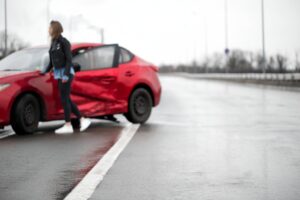 Red car with side impact damage on the side of the highway with a person standing next to the car's door
