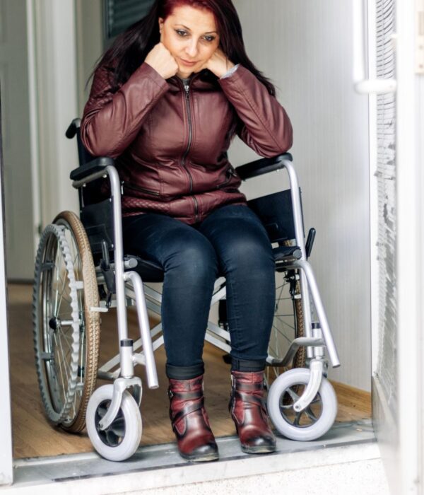 A Baltimore woman sits in a wheelchair at the edge of a doorway, reflecting on her life after suffering a disabling injury from a hit and run car accident. Wearing a burgundy jacket and boots, she appears emotionally impacted by the event that led to her long-term condition. Baltimore Hit and Run Car Accident Lawyers at Regan Zambri Long PLLC help victims like her pursue justice and compensation for permanent injuries. Contact the firm at (202) 972-3767 for experienced legal representation.